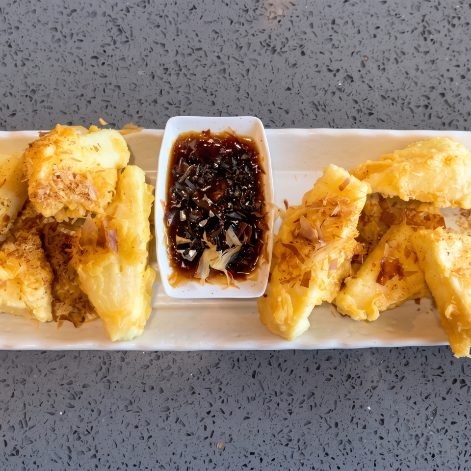 Crispy fried tofu pieces with bonito flakes and a side of dark dipping sauce on a white platter.