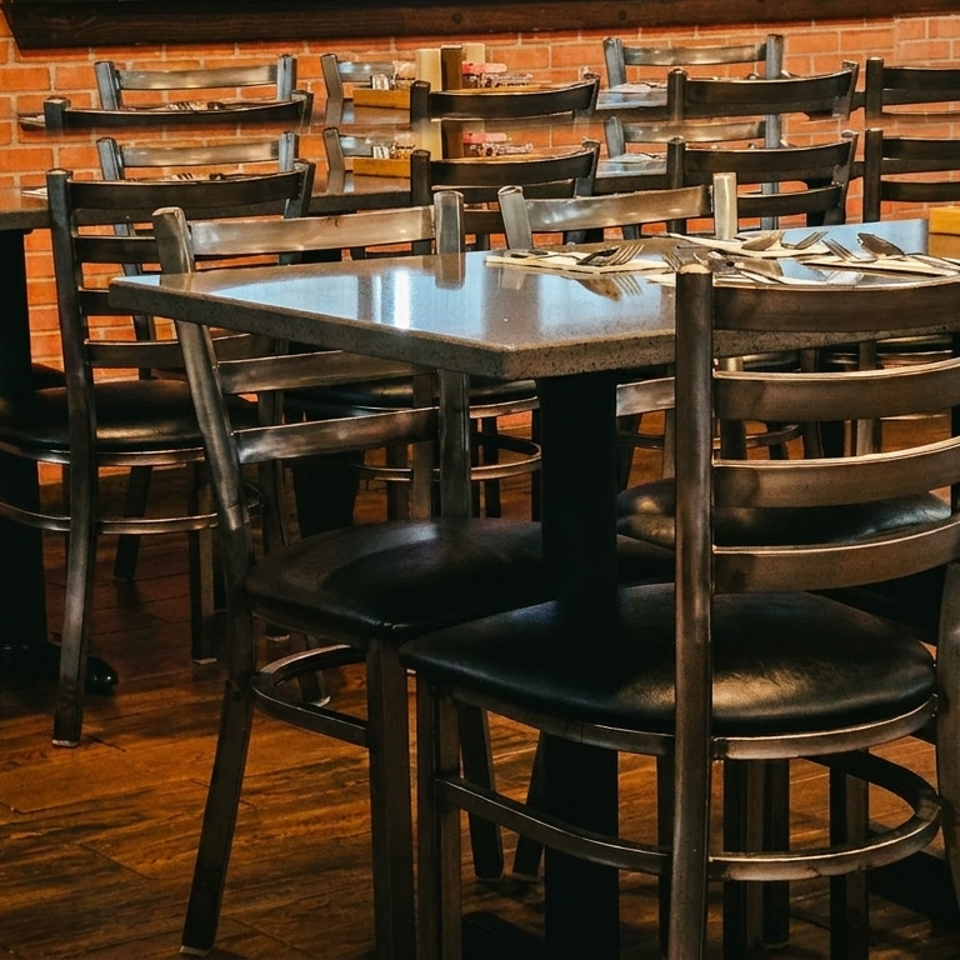 Empty restaurant dining room with dark tables and chairs against a brick wall.