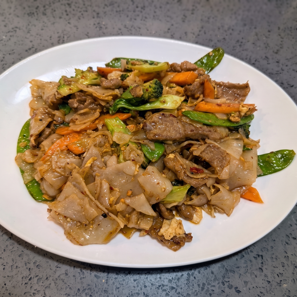 A white plate filled with Pad See Ew, featuring wide noodles, beef, broccoli, carrots, and snow peas.