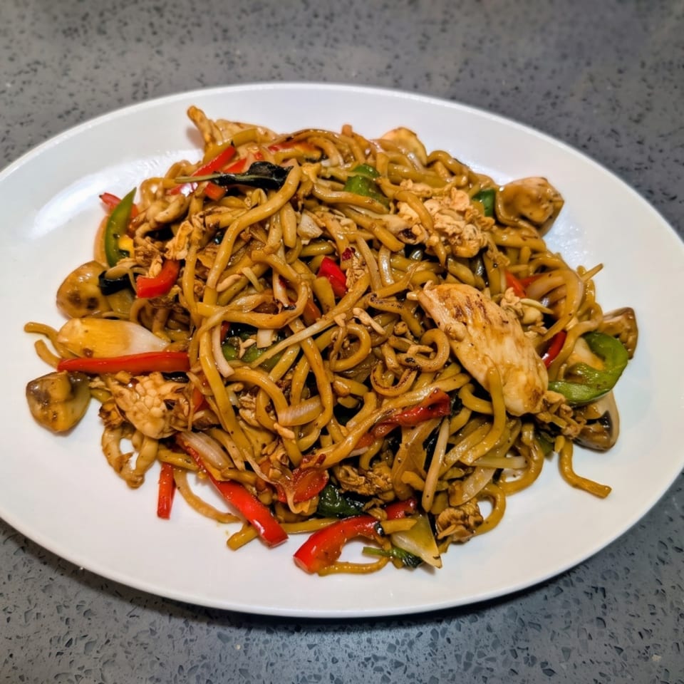 A white plate filled with chicken lo mein, featuring noodles, chicken, and colorful vegetables.